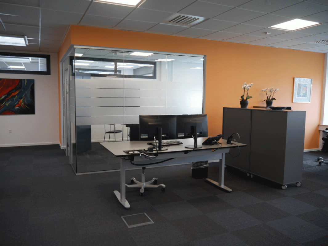 kontorindretning med mørkt ternet gulv, orange væg, gråt arbejdsbord med fladskærme og grå arkivskabe, samt glasrude til kontor i baggrunden.