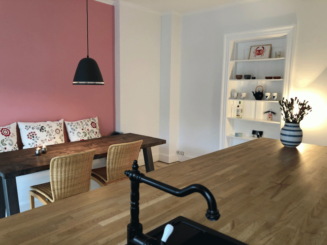 Stueindretning med feng shui. trætema med træborde, træstole, køkkenbord af træ og lyserød væg med farverige puder og sort lampe, samt hvid reol og stribet vase.