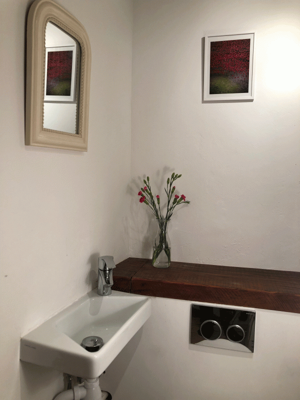 hvidt badeværelse med hvid vask, samt træhylde med blomster, og et billede med lyserøde træer og et spejl på hvid væg.